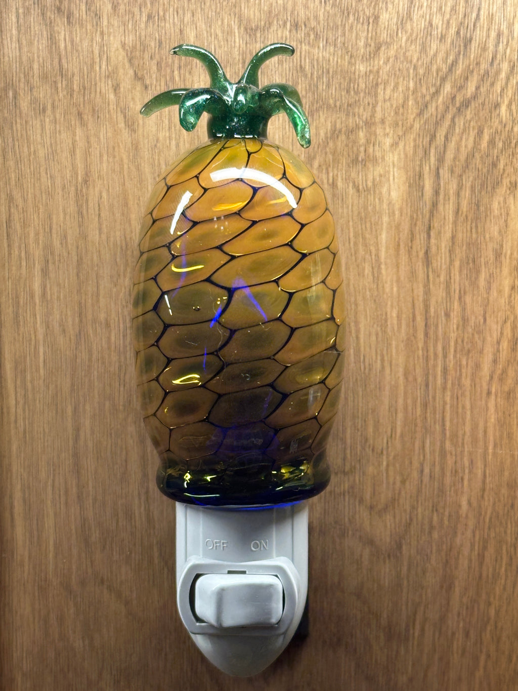 A flameworked and hand-spun blown glass pineapple night light featuring a detailed green crown and a textured yellow frit body that diffuses a warm, amber glow.