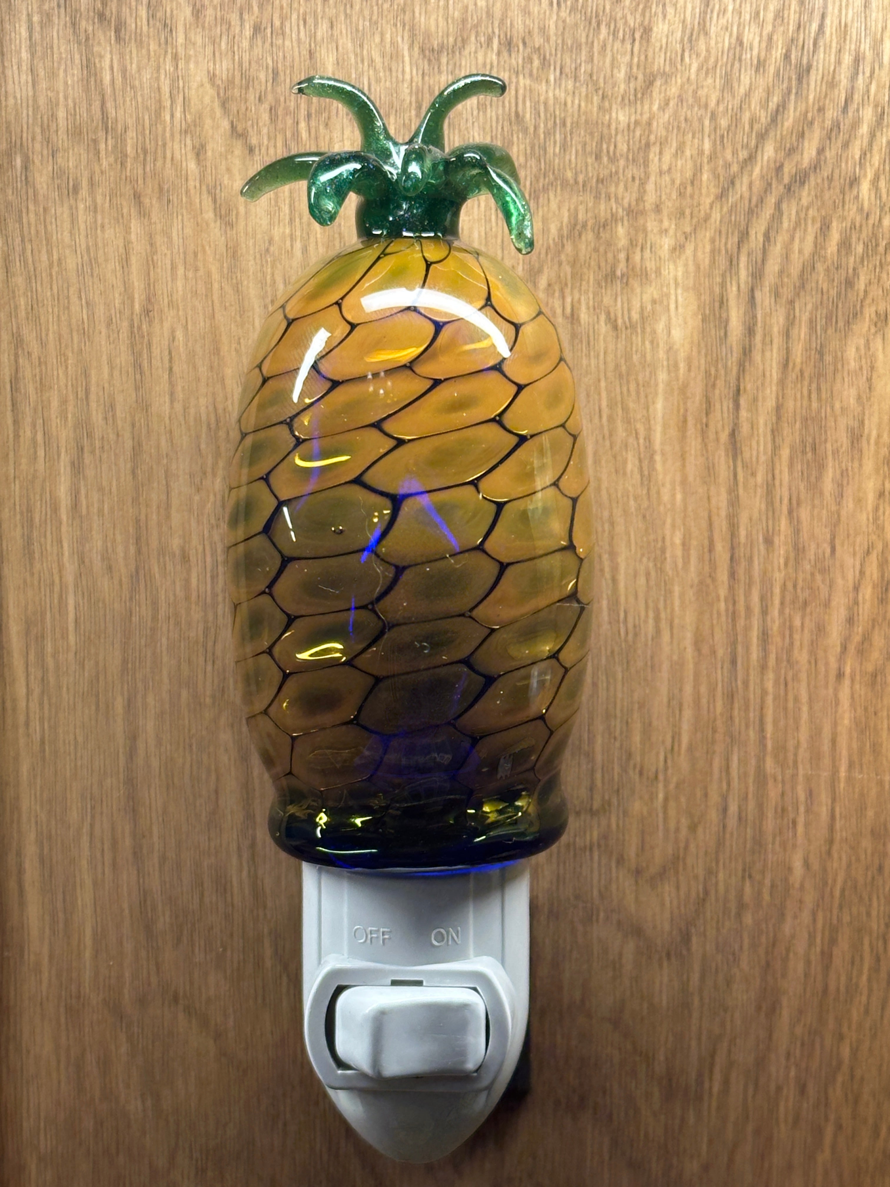 A flameworked and hand-spun blown glass pineapple night light featuring a detailed green crown and a textured yellow frit body that diffuses a warm, amber glow.