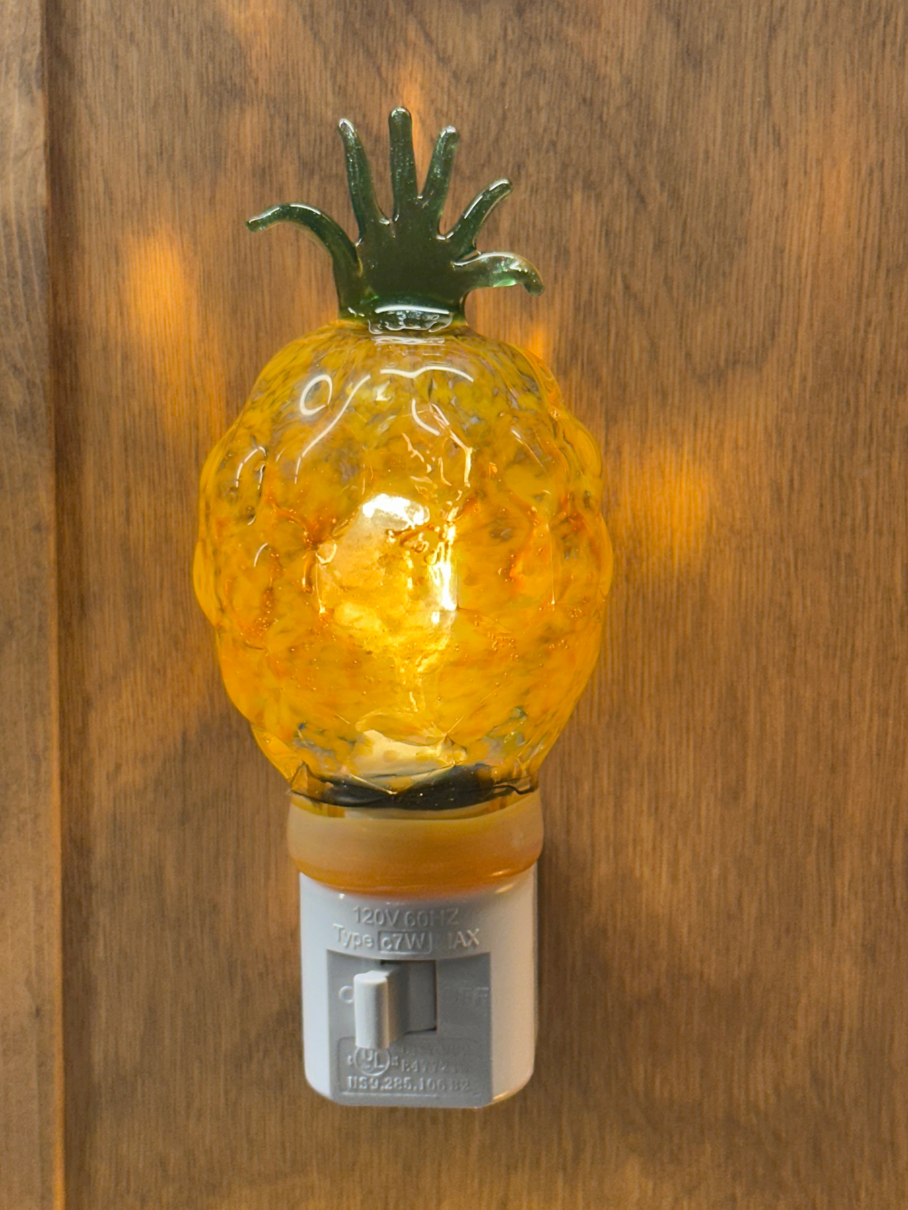 A flameworked and hand-spun blown glass pineapple night light featuring a detailed green crown and a textured yellow frit body that diffuses a warm, amber glow.