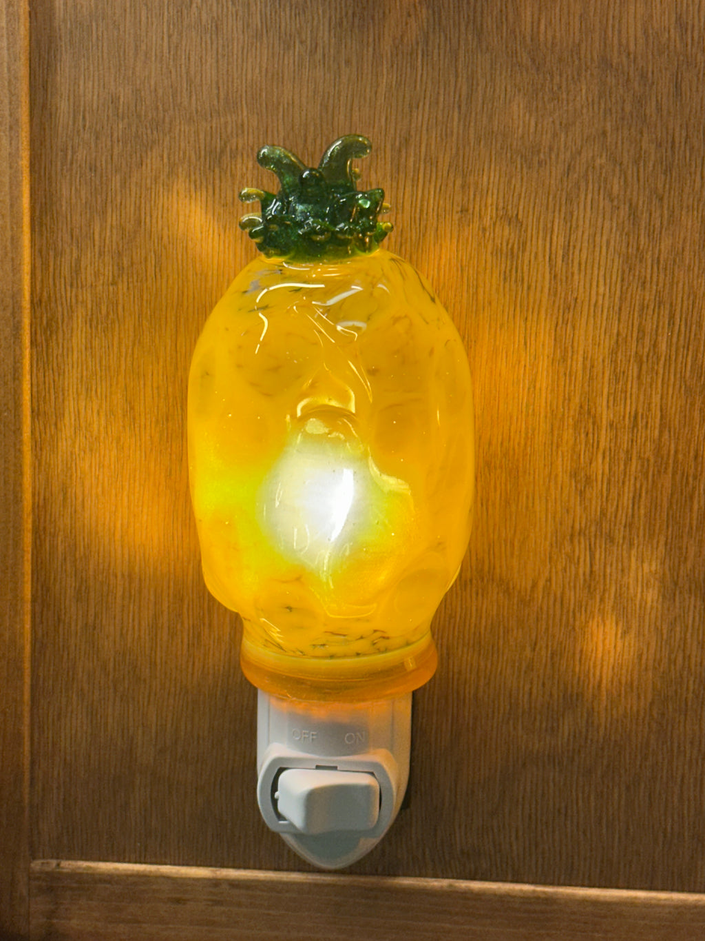 A flameworked and hand-spun blown glass pineapple night light featuring a detailed green crown and a textured yellow frit body that diffuses a warm, amber glow.