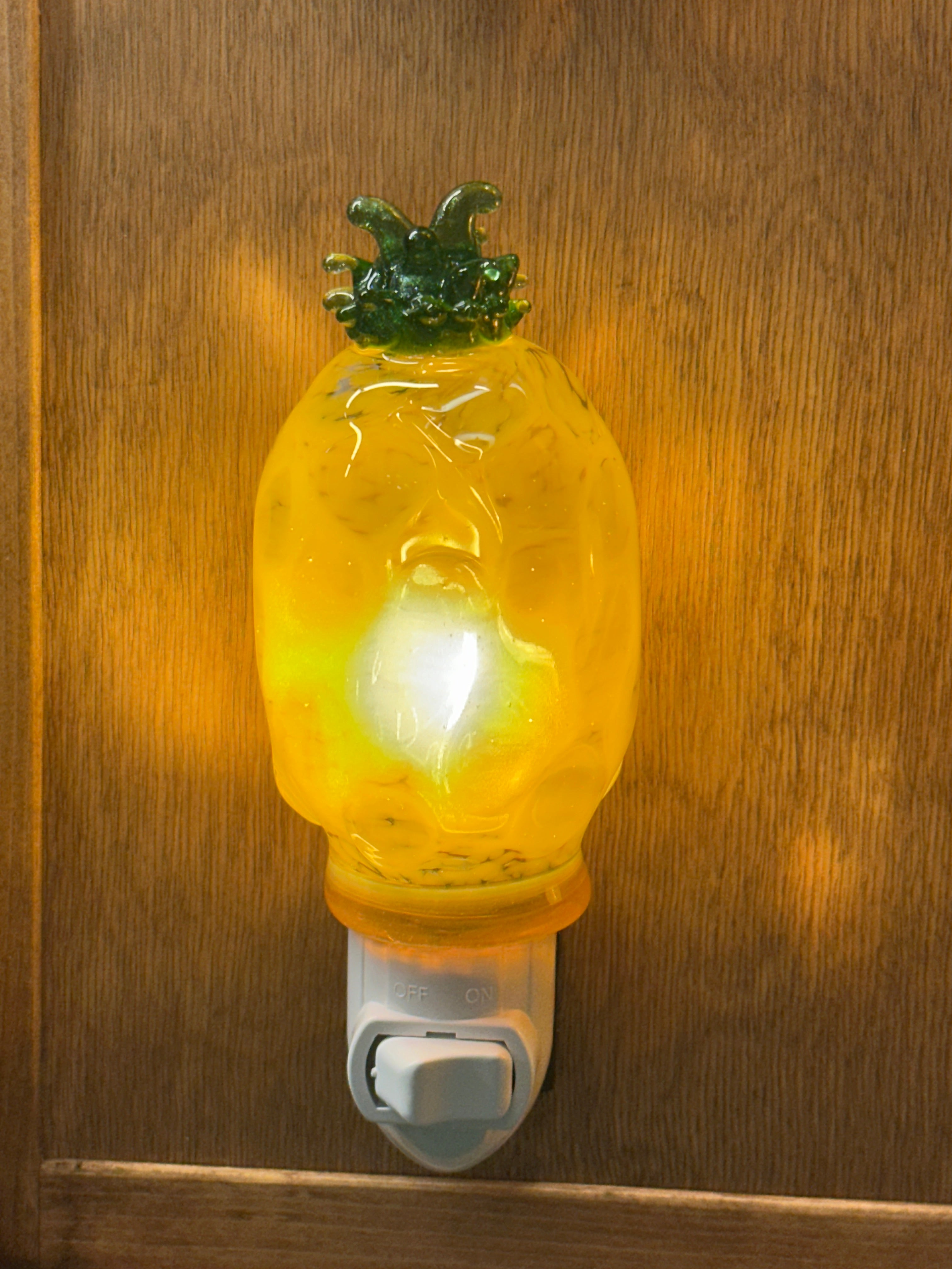A flameworked and hand-spun blown glass pineapple night light featuring a detailed green crown and a textured yellow frit body that diffuses a warm, amber glow.