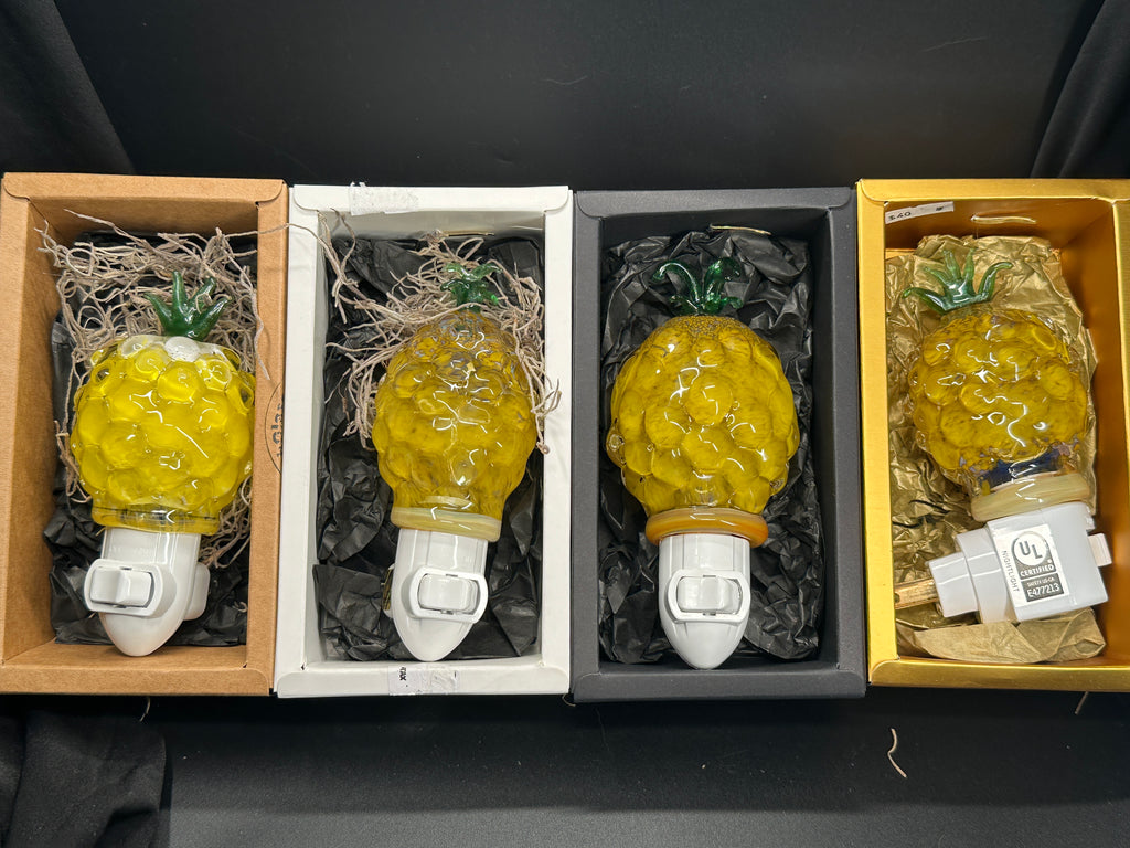 A flameworked and hand-spun blown glass pineapple night light featuring a detailed green crown and a textured yellow frit body that diffuses a warm, amber glow.