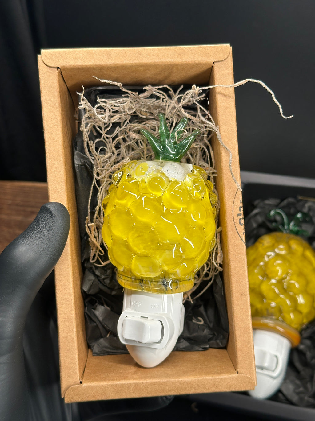 A flameworked and hand-spun blown glass pineapple night light featuring a detailed green crown and a textured yellow frit body that diffuses a warm, amber glow.