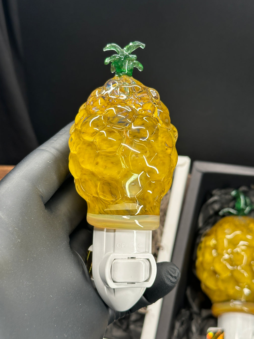 A flameworked and hand-spun blown glass pineapple night light featuring a detailed green crown and a textured yellow frit body that diffuses a warm, amber glow.