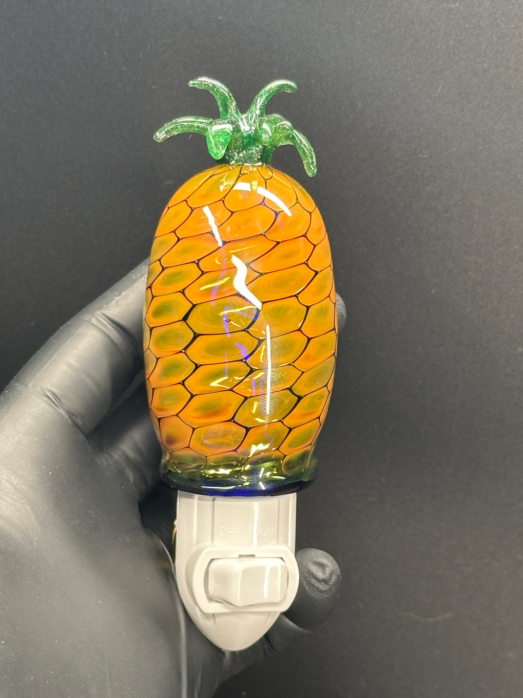 A flameworked and hand-spun blown glass pineapple night light featuring a detailed green crown and a textured yellow frit body that diffuses a warm, amber glow.