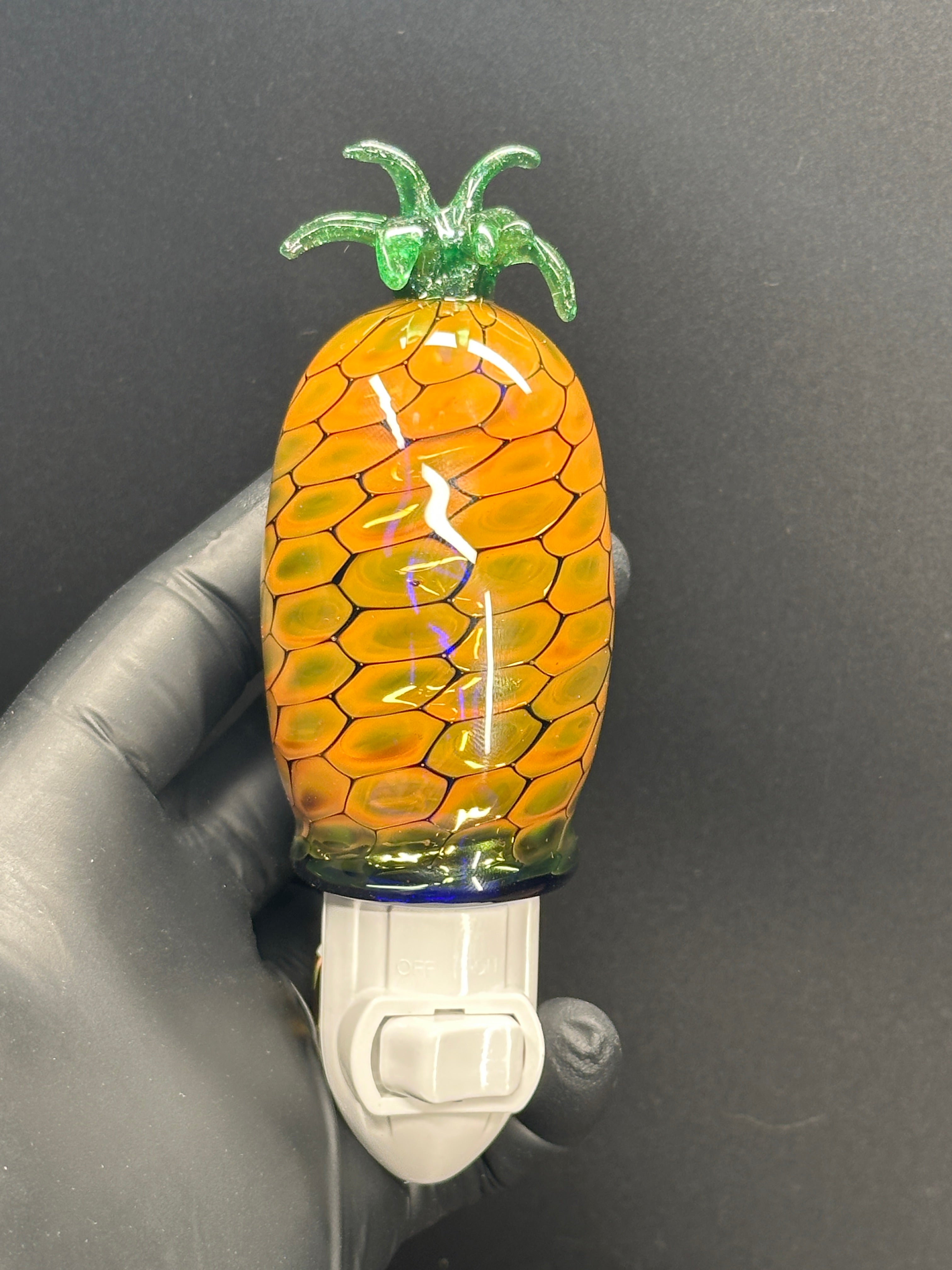 A flameworked and hand-spun blown glass pineapple night light featuring a detailed green crown and a textured yellow frit body that diffuses a warm, amber glow.