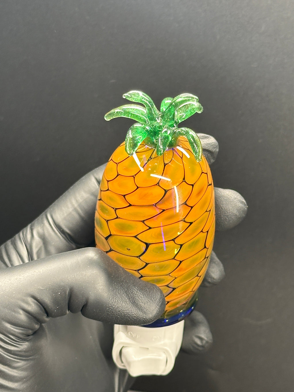 A flameworked and hand-spun blown glass pineapple night light featuring a detailed green crown and a textured yellow frit body that diffuses a warm, amber glow.