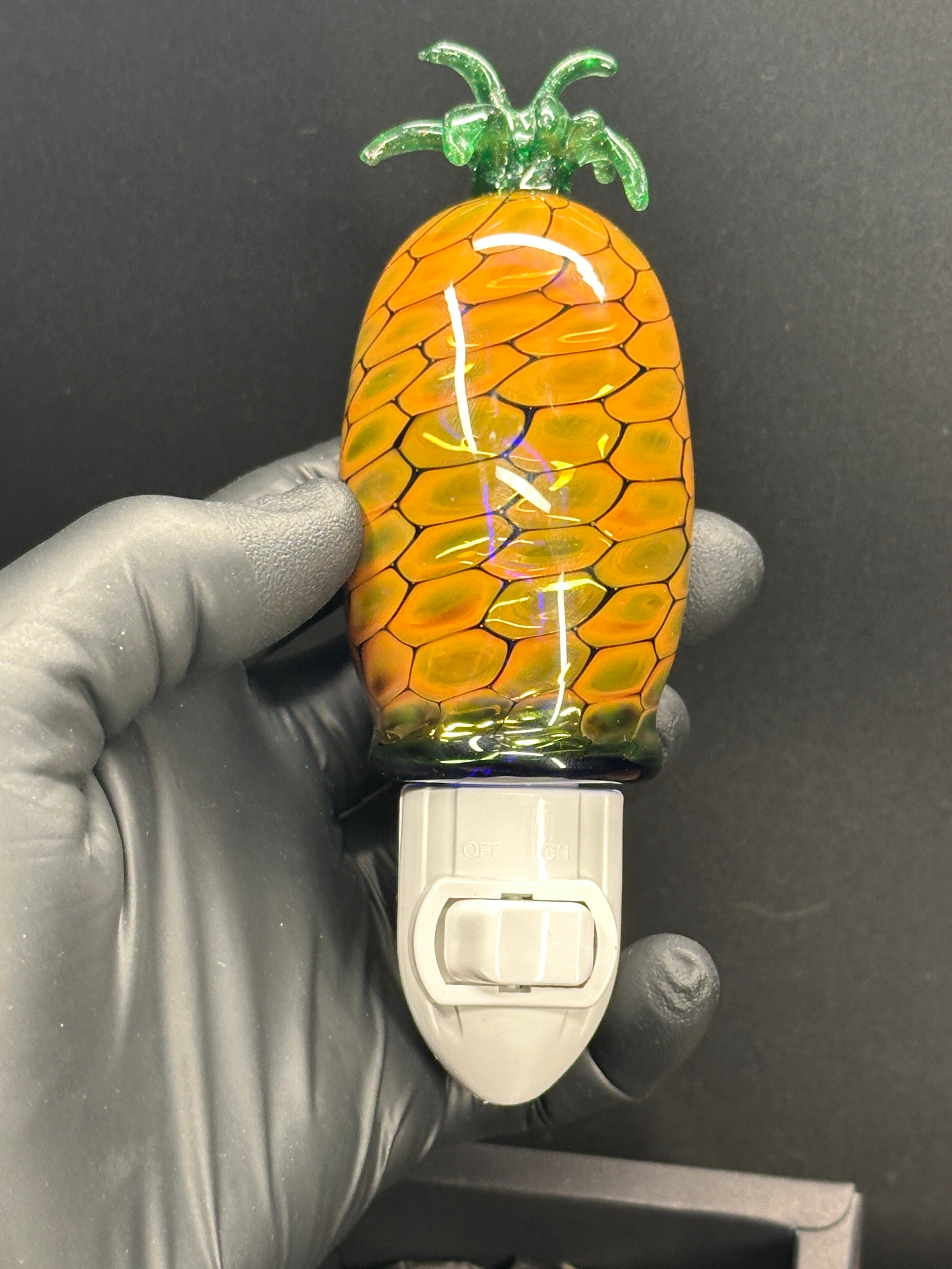 A flameworked and hand-spun blown glass pineapple night light featuring a detailed green crown and a textured yellow frit body that diffuses a warm, amber glow.
