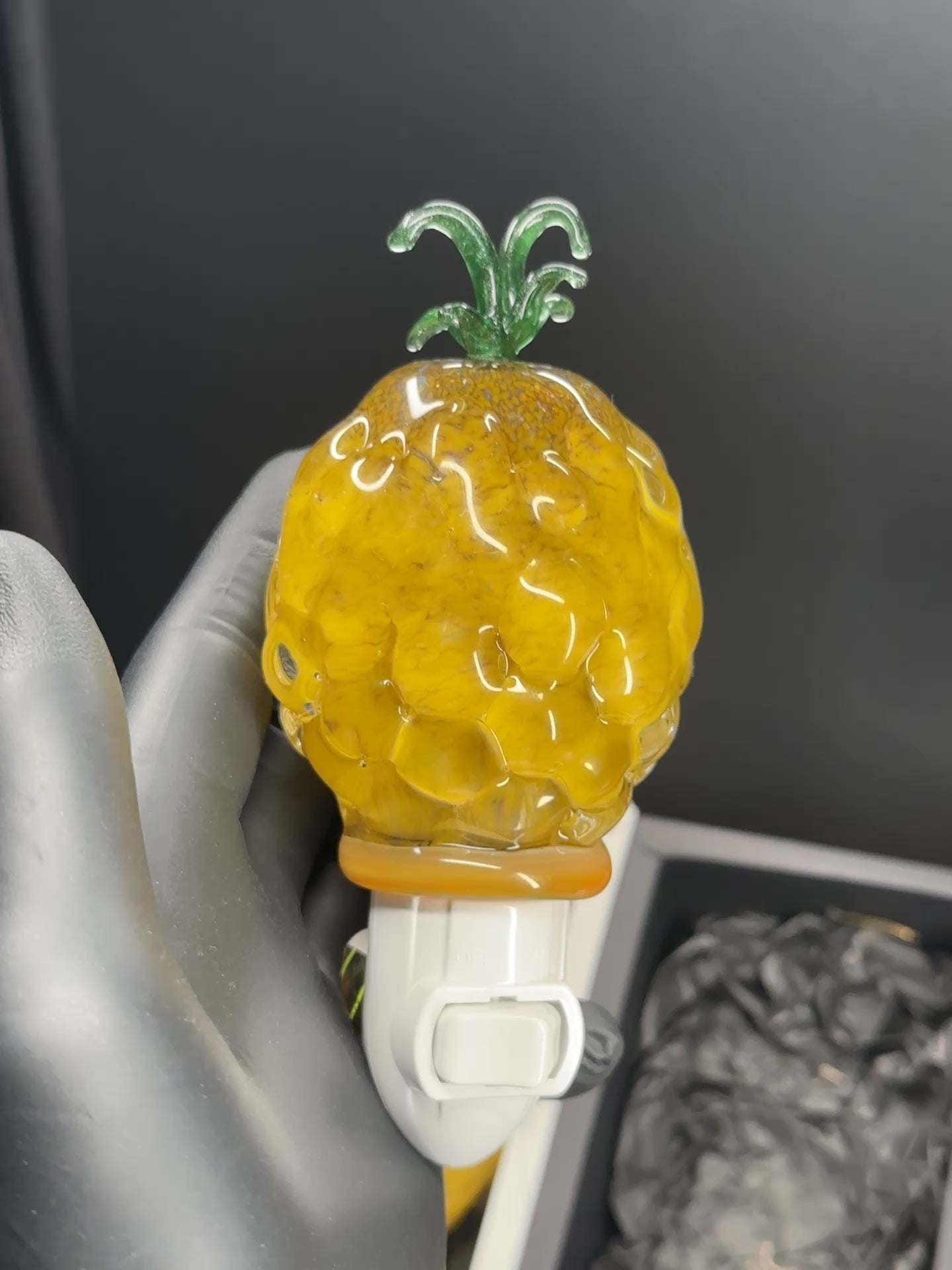 A flameworked and hand-spun blown glass pineapple night light featuring a detailed green crown and a textured yellow frit body that diffuses a warm, amber glow.