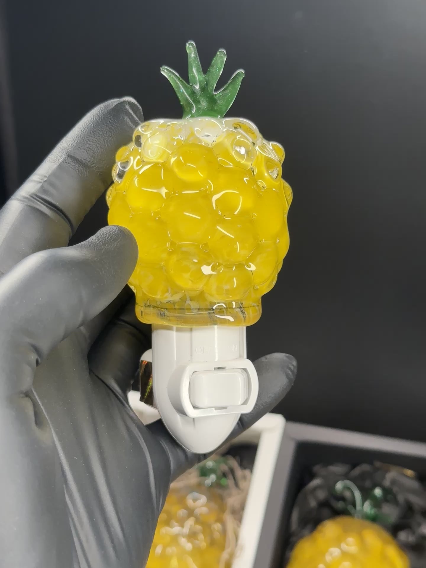 A flameworked and hand-spun blown glass pineapple night light featuring a detailed green crown and a textured yellow frit body that diffuses a warm, amber glow.